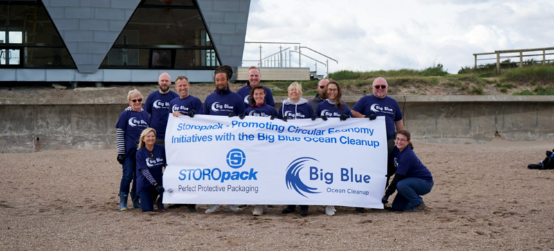 Mehrere Menschen sind zu einem Gruppenfoto am Strand aufgestellt und halten einen Banner des Big Blue Ocean Cleanup.